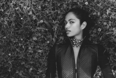 Model lying among dense ground foliage wearing a quilted dark zip jacket and metal statement necklace