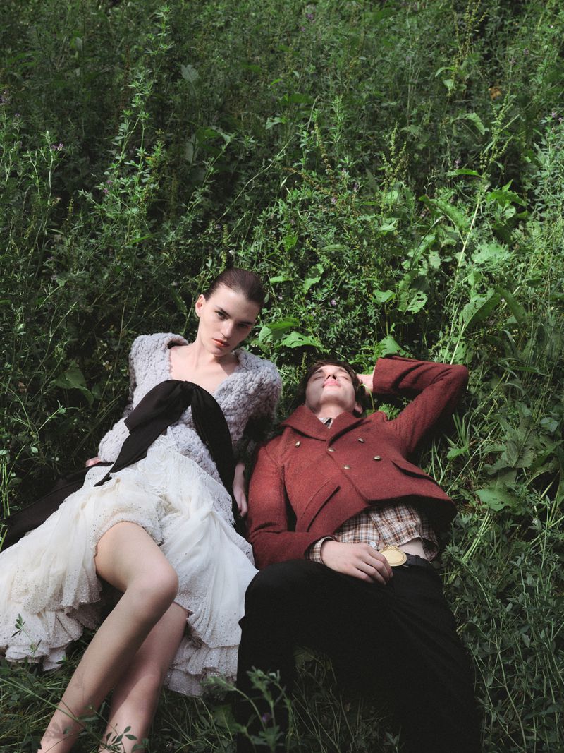 Two models lying in tall green grass, a woman in a white dress and a man in a red jacket