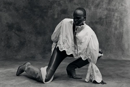 Model Nyibol Dok Jok kneeling in a white embroidered blouse and dark pants against a textured grey background.