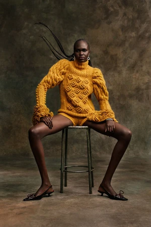 Model Nyibol Dok Jok wearing a yellow cable-knit sweater and shorts seated on a metal stool against a mottled dark backdrop
