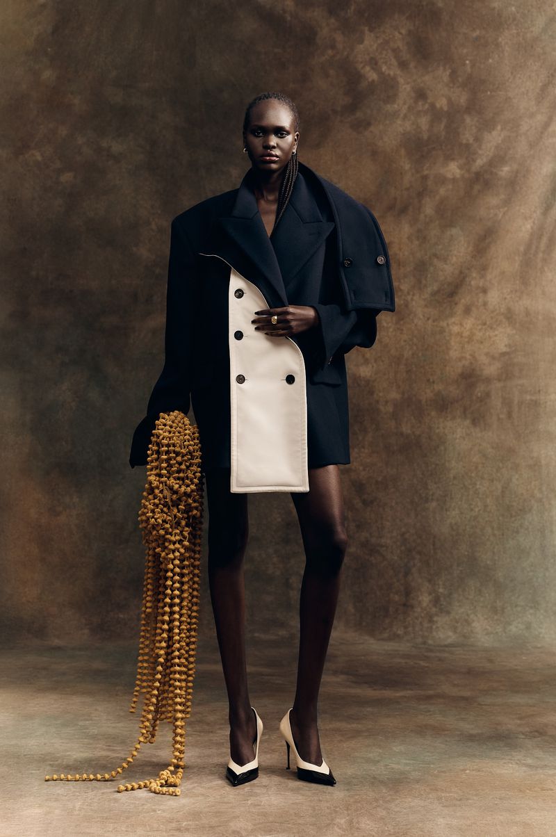 Model Nyibol Dok Jok wearing an oversized dark coat and white heels against a textured backdrop for Glamour Germany.