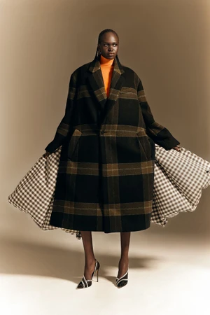 Model Nyibol Dok Jok wearing an oversized plaid coat and black heels against a neutral studio background
