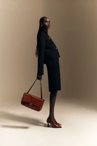 Model Nyibol Dok Jok wearing a black jacket and skirt holding a brown handbag against a neutral studio backdrop for Glamour Germany.