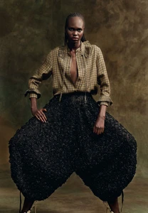 Model Nyibol Dok Jok wearing patterned shirt and textured black trousers against a mottled dark background.
