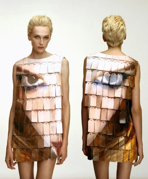 Model Alex Consani wearing a shimmering sequined dress with a printed eye design against a white studio background.