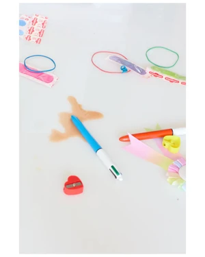 Top-down view of stationery items including a blue pen resting in a liquid spill, a red heart pencil sharpener, and colourful rubber bands.