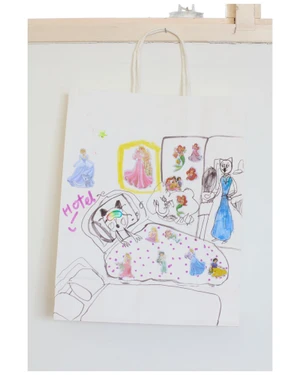 White paper shopping bag with hand-drawn illustrations and princess stickers hanging against a plain white background.
