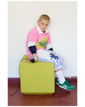 Young child seated on a lime green cube holding a microphone while wearing a pink polo shirt and patterned trousers