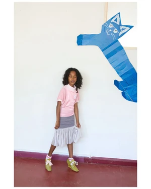Young Black girl in pink top and grey ruffled skirt standing against a white wall with a blue paper cat artwork.