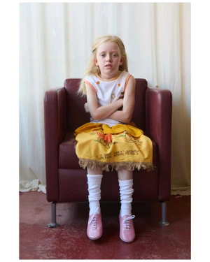 Young child with blonde hair wearing white top, patterned yellow skirt, white socks, and pink shoes seated in maroon armchair
