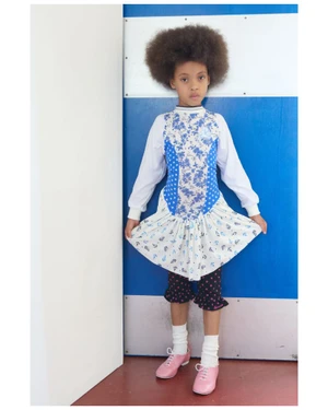 Young child with a large afro hairstyle standing in a patterned dress against a blue and white wall background.