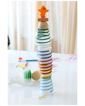 A precariously balanced tall stack of plastic cups, colorful spring toys, wooden spheres, and a small orange plastic figure on top.