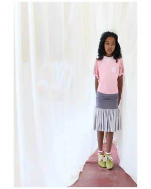 Young child model with dark curly hair wearing a pink top and grey skirt standing against white drapes for i-D Magazine