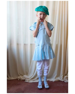 A young child wearing a blue dress, white trousers, blue socks, and a green beret, pulling cheeks in front of a white curtain.
