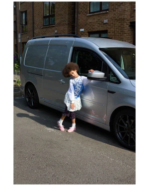 Young child leaning against a parked silver van wearing patterned clothing and pink shoes