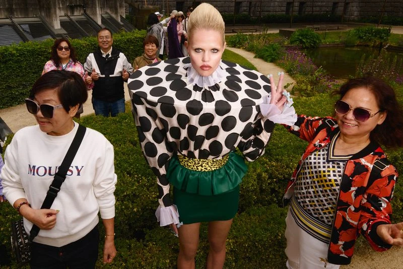 A woman in a polka-dot outfit poses in a garden surrounded by tourists holding up a peace sign in an outdoor, daylight setting.