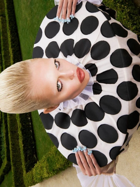 High-angle shot of an adult woman with a blonde hairstyle, wearing a white and black polka dot dress, posing in a garden maze.