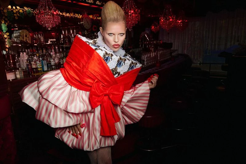Stylish woman in a red and white striped dress poses inside a dimly lit bar with a red chandelier, striking a captivating pose.