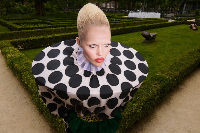 Woman in polka dot dress stands in garden maze. Fashion shot features formal garden with hedge maze and geometric design.