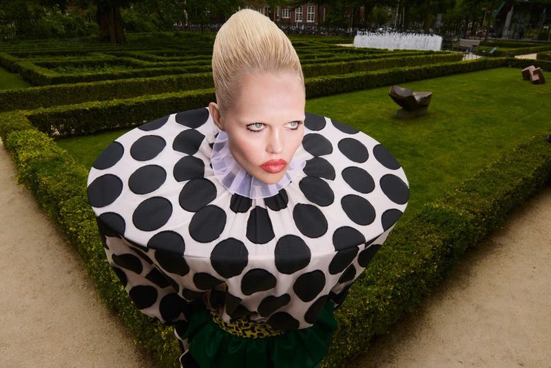 Woman in polka dot dress stands in garden maze. Fashion shot features formal garden with hedge maze and geometric design.