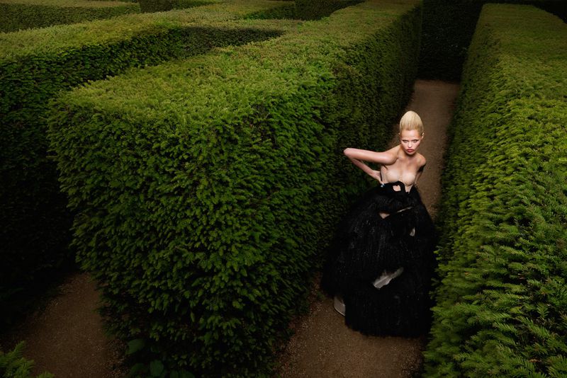 Blonde model poses in black dress within a green hedge maze, captured in medium shot with a shallow depth of field.