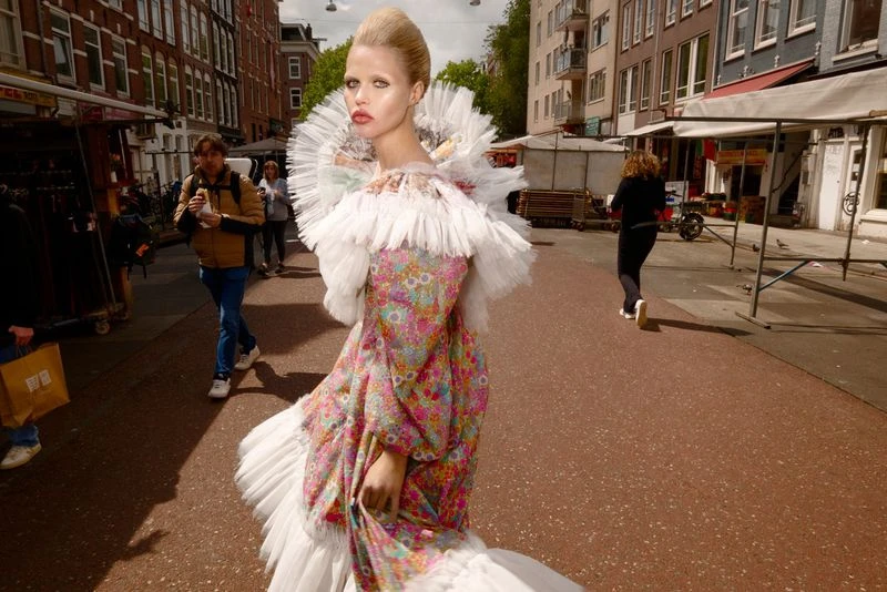 A woman walks down a city street wearing a pink floral gown with a large white ruffled collar in a high fashion editorial shot.