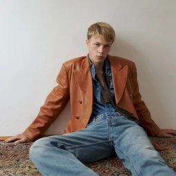 Blonde male model wearing a tan leather blazer and blue denim jeans seated against a plain white wall
