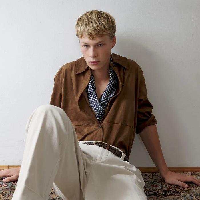 Blonde male model wearing a brown shirt and white trousers seated against a plain wall