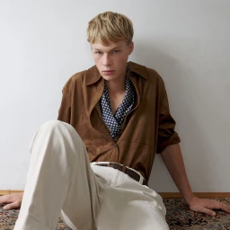 Blonde male model wearing a brown shirt and white trousers seated against a plain wall