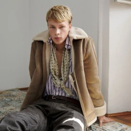 Young male model sitting on a patterned rug wearing a tan shearling jacket and striped shirt for U Repubblica editorial.