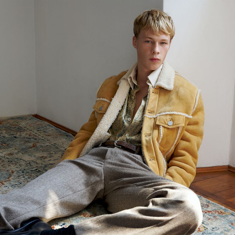 Male model seated on a patterned rug wearing a yellow shearling jacket, patterned shirt, and grey trousers.