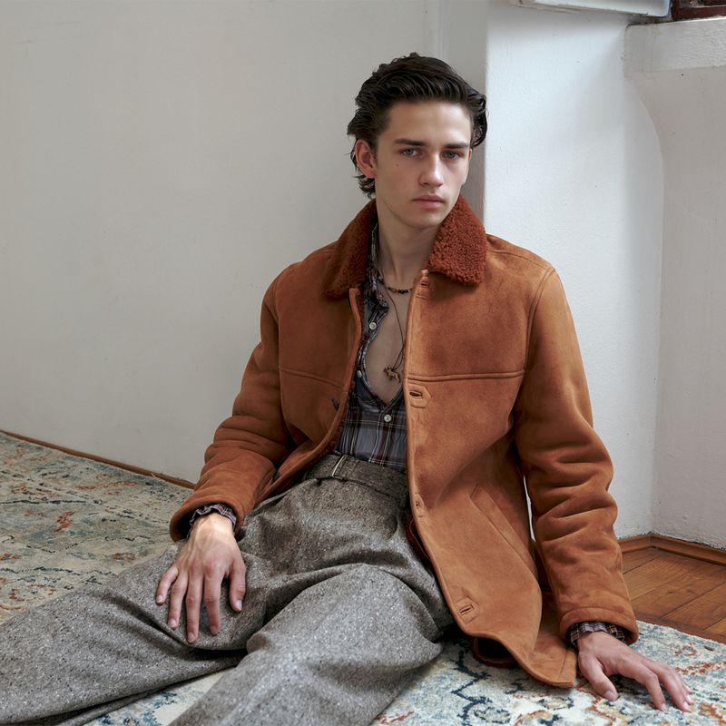Young male model wearing a brown shearling-collar jacket seated on a patterned rug indoors