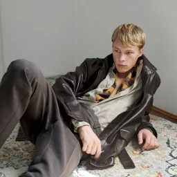 Young male model reclining on patterned rug against plain wall wearing leather jacket and striped shirt
