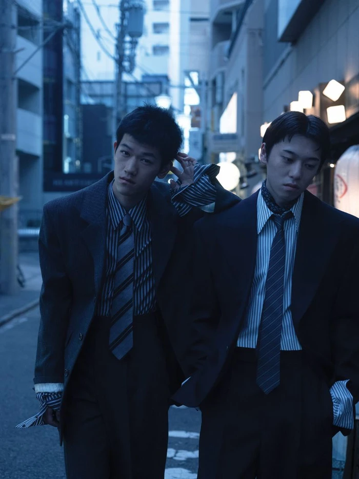 Two young men in tailored suits and striped ties posing in a narrow city street at twilight