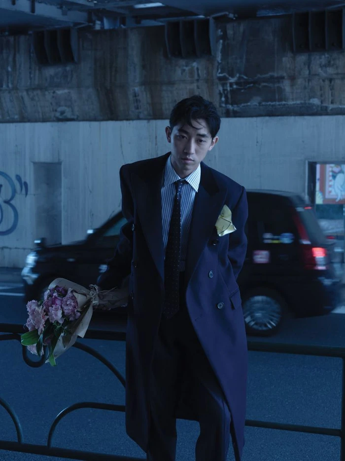 Man wearing a dark double-breasted overcoat and striped shirt holding a bouquet of flowers by a street.