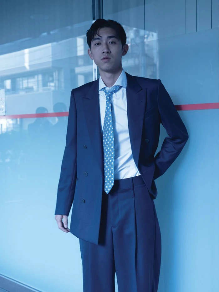 Man wearing a navy blue double-breasted suit and light blue patterned tie against a blue frosted glass wall