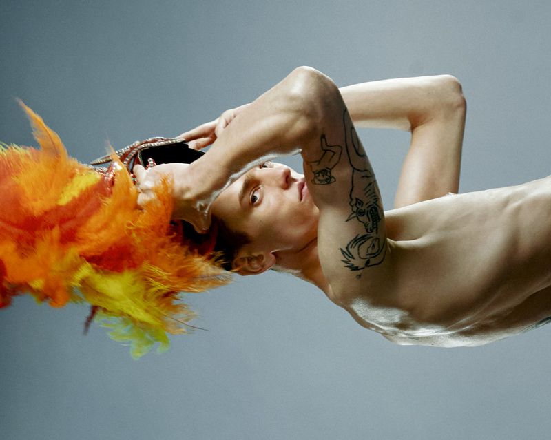Shirtless male model with arm tattoos holds an ornate orange and yellow feathered headpiece against a grey backdrop.