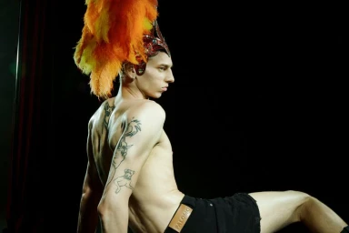 Shirtless male model wearing an orange and yellow feather headdress and black shorts against a dark background