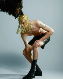 Shirtless male model wearing an ornate gold beaded headdress with black feathers, dark shorts, and socks against a grey background.