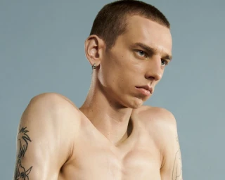 Close-up portrait of a young man with a buzz cut, tattoos on his arms, and a small hoop earring against a light grey background.