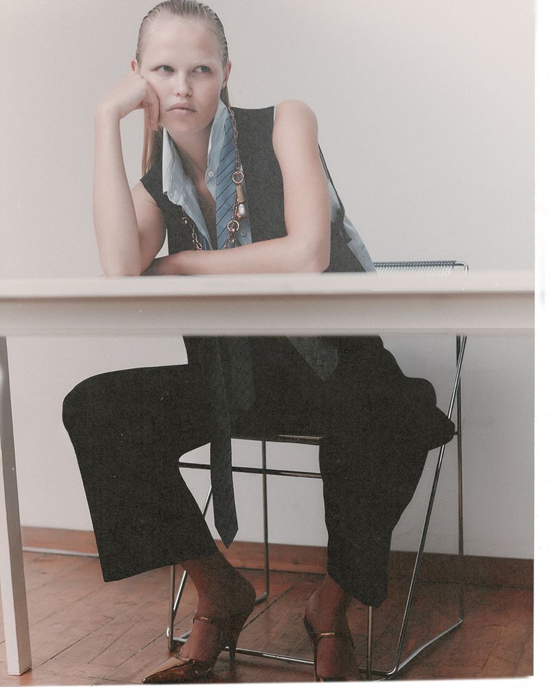 Model seated at a white table wearing a dark vest and trousers with a light blue shirt and tie