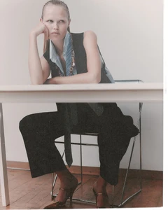 Model seated at a white table wearing a dark vest and trousers with a light blue shirt and tie
