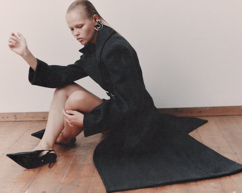Model wearing a dark coat and black heels seated on a wooden floor against a plain wall for WRPD Magazine