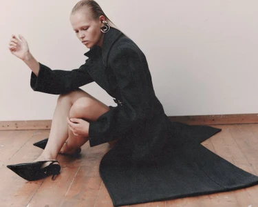 Model wearing a dark coat and black heels seated on a wooden floor against a plain wall for WRPD Magazine