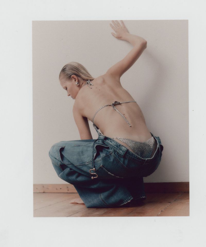 Model squatting against a white wall wearing low-slung denim cargo pants and a sparkling bikini top for WRPD Magazine.