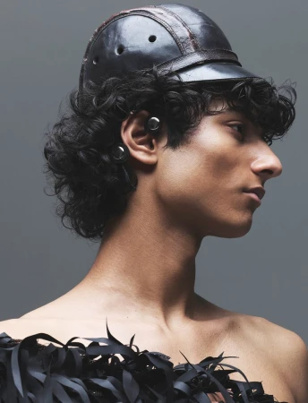 Profile portrait of a male model wearing a dark leather cap and fringe garment against a neutral grey studio backdrop.