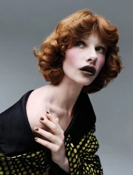 Close-up portrait of a red-haired model in dark lipstick and a patterned black and yellow garment against a grey background.
