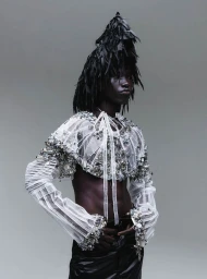 Model in embellished sheer white cropped top with black feathered headpiece and dark trousers against a grey backdrop