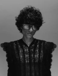 Close-up portrait of a model with dark curly hair wearing oversized sunglasses and a black lace top against a plain grey background.