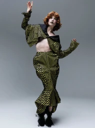 Model with red hair wearing a patterned yellow and black outfit posing against a grey background for The Hunger Magazine